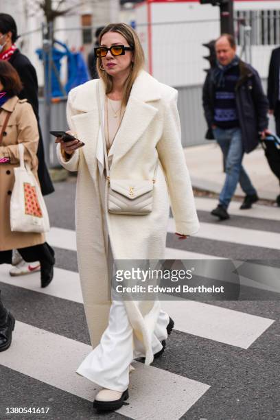 Guest wears a beige t-shirt, a beige long oversized coat, a white latte crossbody bag from Saint Laurent, white flared pants, beige leather with...