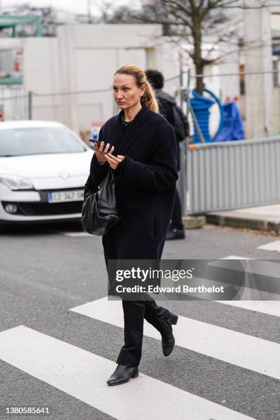 Guest wears a black t-shirt, a black long oversized coat, a black shiny leather handbag, black suit pants, black shiny leather block heels ankle...