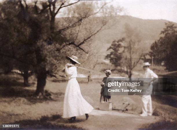 Watched by a male companion and a caddy, a woman in a sundress and hat swings a golf club, early twentieth century.