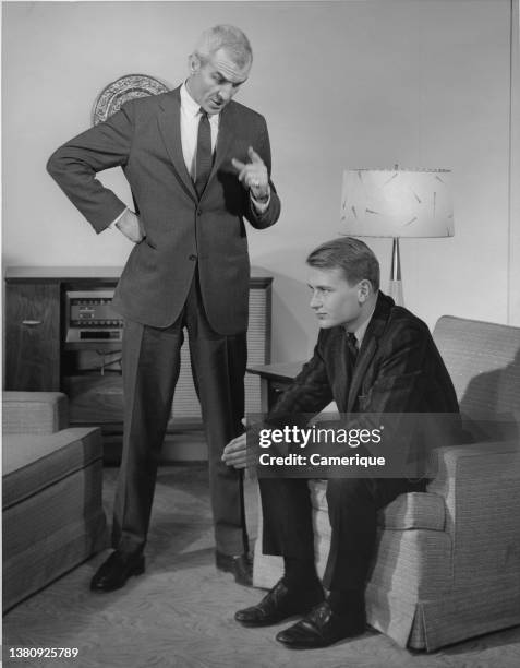 Older dad standing up making hand gestures to teenage son who is sitting in a chair staring straight ahead with hands together.