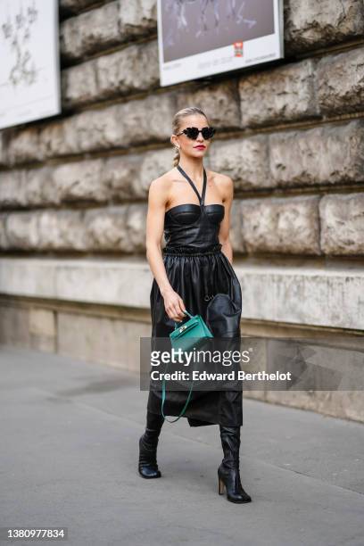Caroline Daur wears black cut-out sunglasses from Loewe, silver and rhinestones earrings, a black shiny leather halter-neck / shoulder-off /...