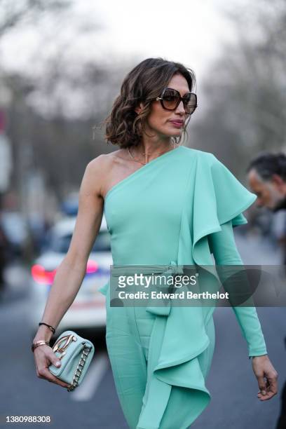Nieves Alvarez wears brown large sunglasses, a green asymmetric shoulder / ruffled shoulder / a long sleeve / belted jumpsuit, gold rings, a black...