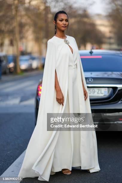 Lais Ribeiro wears diamond earrings, a white long cloak from Elie Saab, a white flowing sleeveless / belted flared jumpsuit from Elie Saab, silver...
