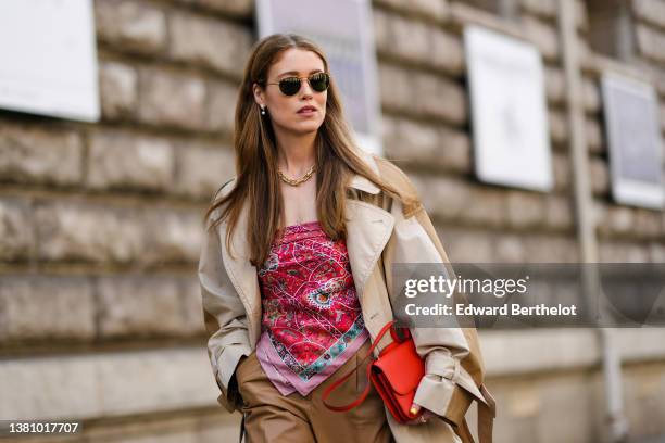 Annabel Rosendahl wears sunglasses, pearls pendant earrings, a gold large chain necklace, a beige and brown long oversized coat, a red shiny leather...