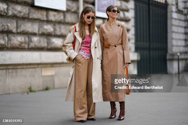 Annabel Rosendahl wears sunglasses, pearls pendant earrings, a gold large chain necklace, a beige and brown long oversized coat, a red shiny leather...