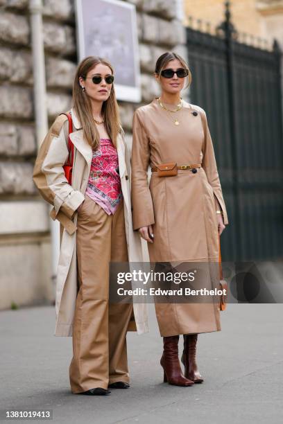 Annabel Rosendahl wears sunglasses, pearls pendant earrings, a gold large chain necklace, a beige and brown long oversized coat, a red shiny leather...