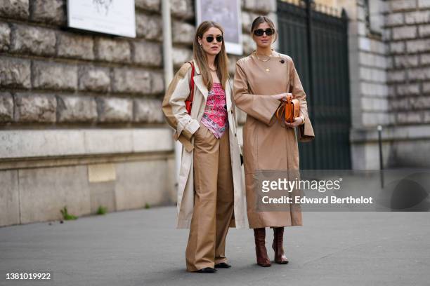 Annabel Rosendahl wears sunglasses, pearls pendant earrings, a gold large chain necklace, a beige and brown long oversized coat, a red shiny leather...