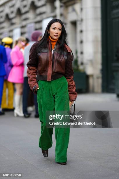 Yoyo Cao wears an orange ribbed wool turtleneck pullover, a brown shiny leather / black leather straps / quilted sleeves zipper jacket, green large...