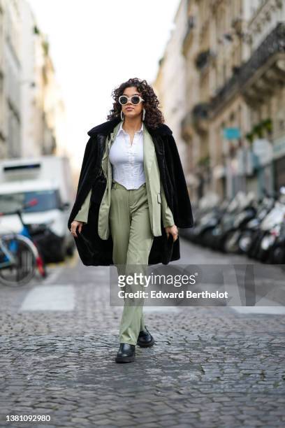 Guest wears white sunglasses, silver earrings, a white corset cropped shirt, a black fluffy coat, a green blazer jacket, matching green suit pants, a...