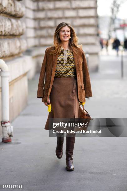 Guest wears gold earrings, a brown suede jacket, a yellow / black / gray print pattern polo shirt, a dark brown wool slit / split knees skirt, a...