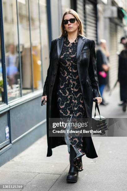 Guest wears black sunglasses, gold earrings, a black with red and green flower print pattern shirt long dress, a black shiny leather long coat, a...