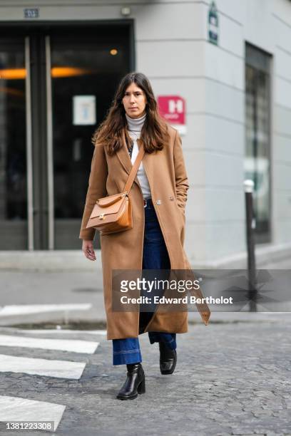 Guest wears a white turtleneck pullover, a brown long wool coat, a brown shiny leather belt, a camel shiny leather crossbody bag, navy blue denim...