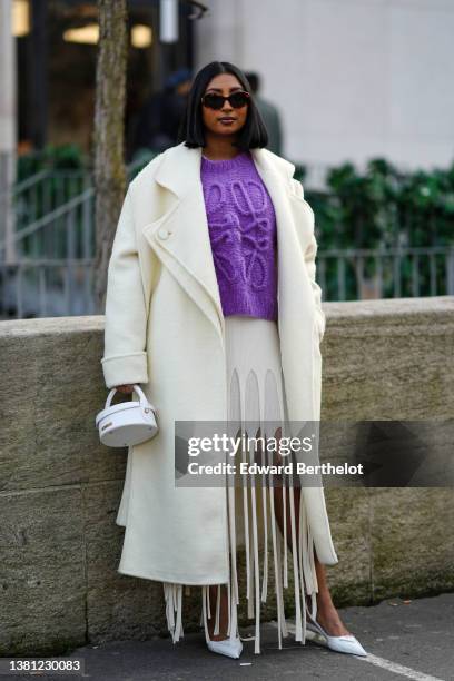 Guest wears black sunglasses, a purple with oversized logo embossed pattern pullover from Loewe, a white latte long oversized coat, a white fringed...