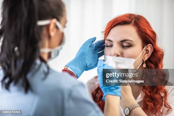 red-haired woman with face mask pulled under her nose lets the doctor to take swab test, closing her eyes. - coronavirus testing stock-fotos und bilder