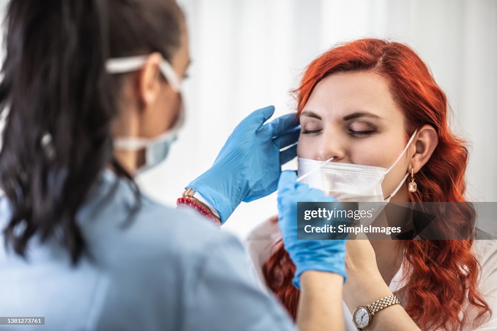 Red-haired woman with face mask pulled under her nose lets the doctor to take swab test, closing her eyes.