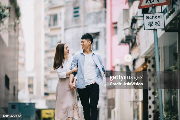 young asian couple holding hands and smiling as they walk through local city street in hong kong, enjoying city life. love and dating concept - stadscentrum hongkong stockfoto's en -beelden