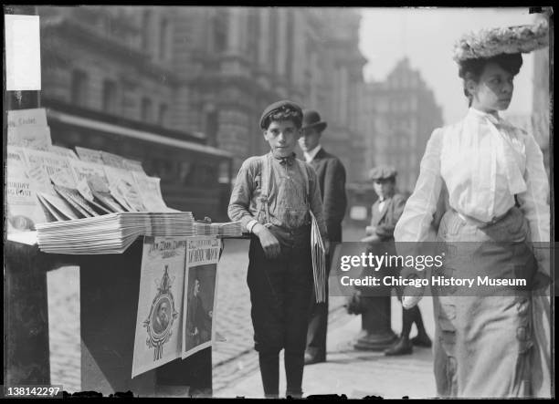 Newsboy holding newspapers and standing next to a paper stand on a commercial street, Chicago, Illinois, August 11, 1904.