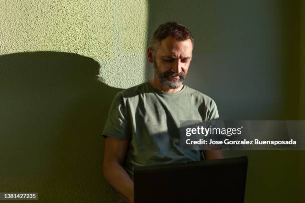 lächelnder mann mit grünem t-shirt auf grünem hintergrund, der mit seinem laptop arbeitet. natürliche lichtseite - schattig stock-fotos und bilder