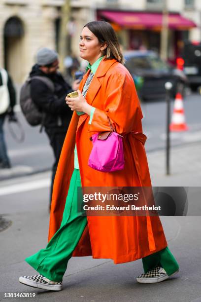 Guest wears gold earrings, a green shirt, a black and white checkered print pattern pullover, a neon orange shiny leather long oversized coat, a pink...