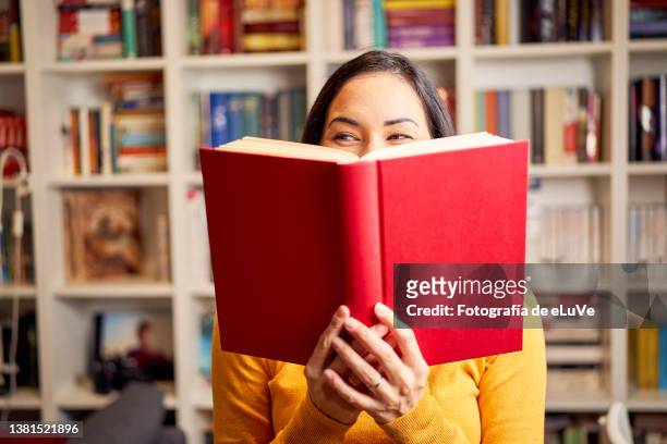female young behind book with face covered for a red book while smiling - reading stock-fotos und bilder