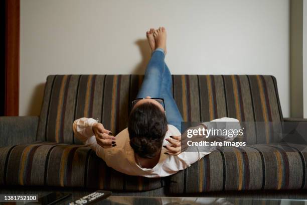 back view of woman doing sit-ups on the couch in her apartment. woman wearing glasses lying on sofa with legs up while exercising. - lying on back ストックフォトと画像