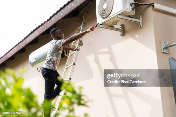 young man standing on ladder stretching dangerously with tool to fix air conditioner - ladder leaning stock pictures, royalty-free photos & images