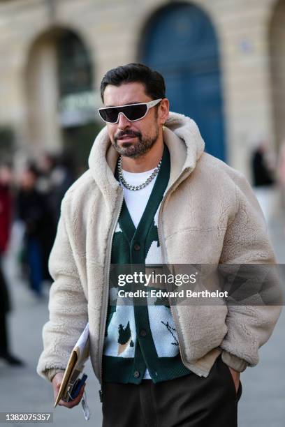 Alex Badia wears white sunglasses from Celine, a gold cross pendant earrings, a white t-shirt, a silver large chain necklace, a dark green / pale...