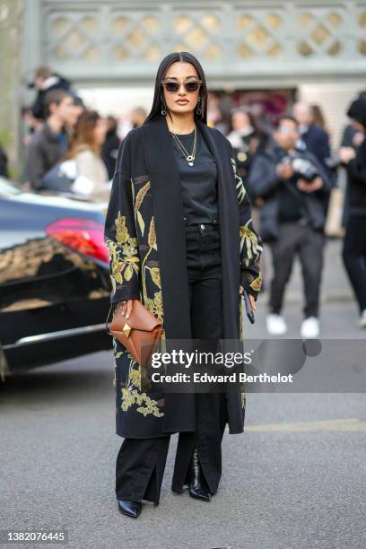 Amina Muaddi wears sunglasses, gold earrings, gold chain pendant necklace, a black t-shirt, a black with green embroidered flower pattern long...