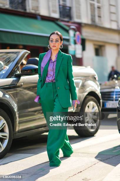 Yoyo Cao wears silver earrings, gold sunglasses, a purple shirt, a dark green blazer jacket, dark green matching large pants, silver rings, pumps...
