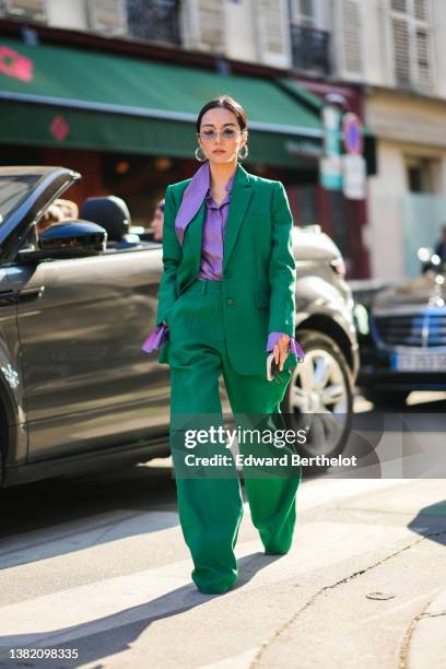 Yoyo Cao wears silver earrings, gold sunglasses, a purple shirt, a dark green blazer jacket, dark green matching large pants, silver rings, pumps...