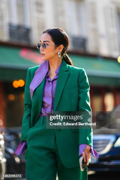 Yoyo Cao wears silver earrings, gold sunglasses, a purple shirt, a dark green blazer jacket, dark green matching large pants, silver rings, outside...