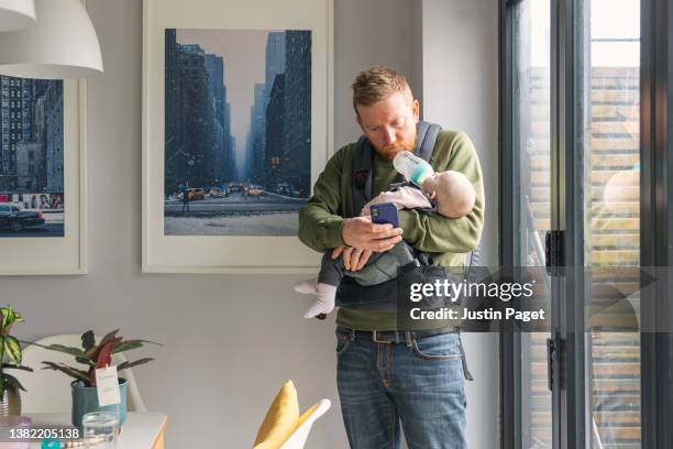 mature man using his phone whilst feeding his baby daughter. he uses his chin to support the baby bottle - multitasking! - multi tasking stock-fotos und bilder