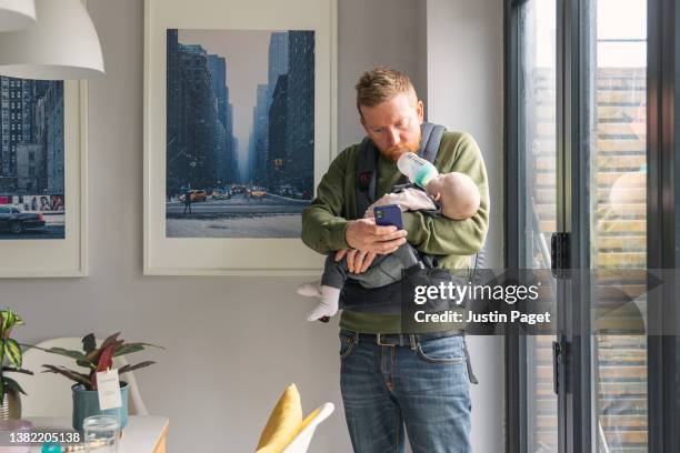 mature man using his phone whilst feeding his baby daughter. he uses his chin to support the baby bottle - multitasking! - new life stock pictures, royalty-free photos & images