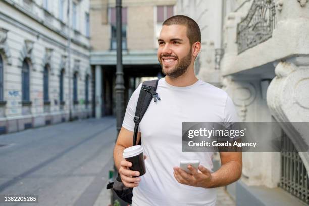 happy young man strolling the city with a phone and coffee to go in his hands - takeaway coffee stockfoto's en -beelden