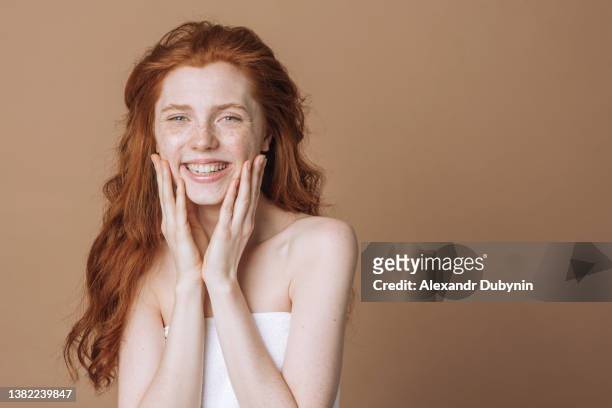 happy young redhead woman with clear skin touching her face with freckles on beige background in studio .smile lady face portrait with copy space. cosmetics and skin care concept - cheveux roux photos et images de collection