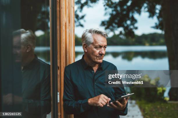 senior man with tablet pc standing by glass wall at backyard - rich man stock-fotos und bilder