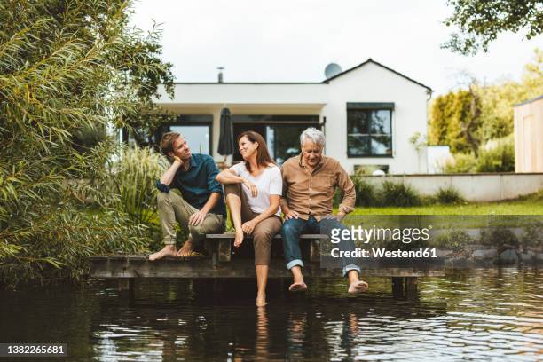 mother and son talking by senior man sitting on jetty by lake at backyard - standing water foto e immagini stock