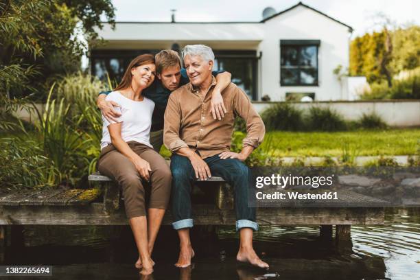 son embracing father and mother sitting on jetty by lake at backyard - bootssteg stock-fotos und bilder