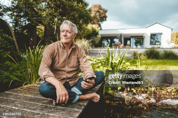 senior man with smart phone sitting cross-legged on jetty by lake - jetty lake stock pictures, royalty-free photos & images
