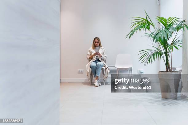 young woman using mobile phone sitting in waiting room at clinic - hospital waiting room stockfoto's en -beelden