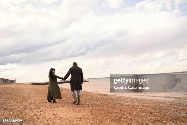 couple in winter coats holding hands walking on ocean beach - romantic couple walking winter beach stock pictures, royalty-free photos & images