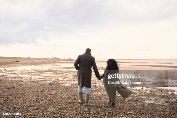 couple in winter coats holding hands walking on ocean beach - romantic couple walking winter beach stock-fotos und bilder