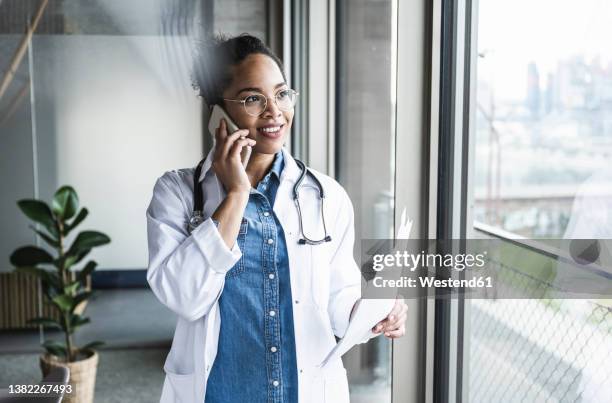 smiling doctor with document talking on smart phone at hospital - healthcare professional stock pictures, royalty-free photos & images