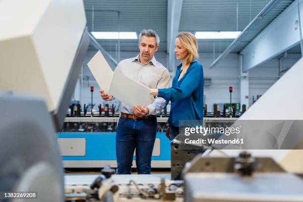 blond businesswoman discussing over report with businessman in factory - papierwerk stock-fotos und bilder