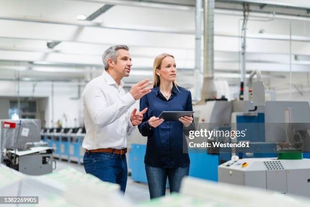 businessman sharing ideas with businesswoman holding tablet pc in factory - 40 44 years blond hair mature man factory stock pictures, royalty-free photos & images