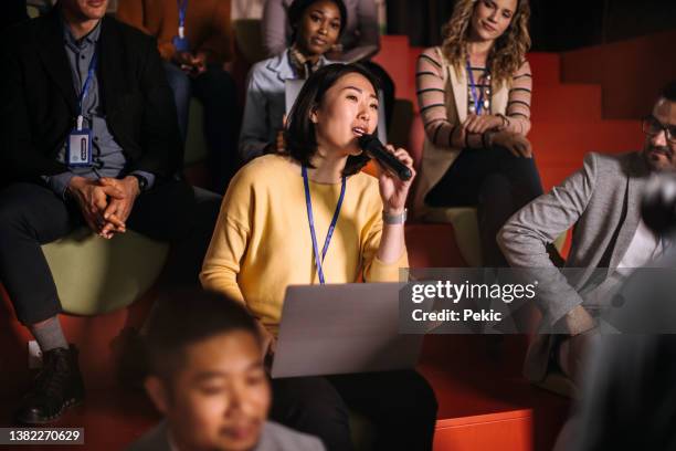 young beautiful asian woman asking a question while attending business conference in a room full of audience - business conference stock pictures, royalty-free photos & images