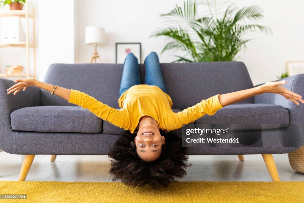 Happy young woman lying upside down on sofa in living room at home