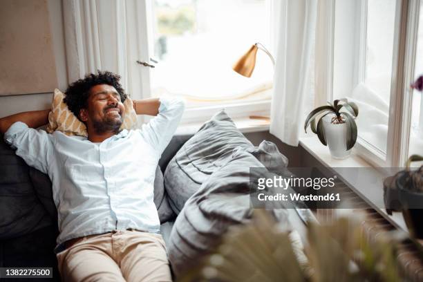smiling man with eyes closed resting on sofa at home - resting imagens e fotografias de stock