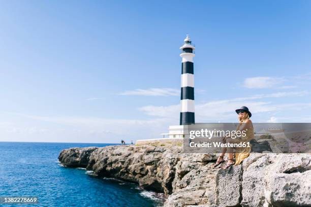 woman sitting on rock by sea at artrutx lighthouse in minorca, spain - menorca stockfoto's en -beelden
