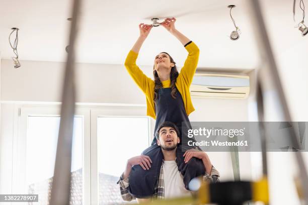 young woman sitting on boyfriend shoulders installing light bulb in new home - ceiling lamp stockfoto's en -beelden